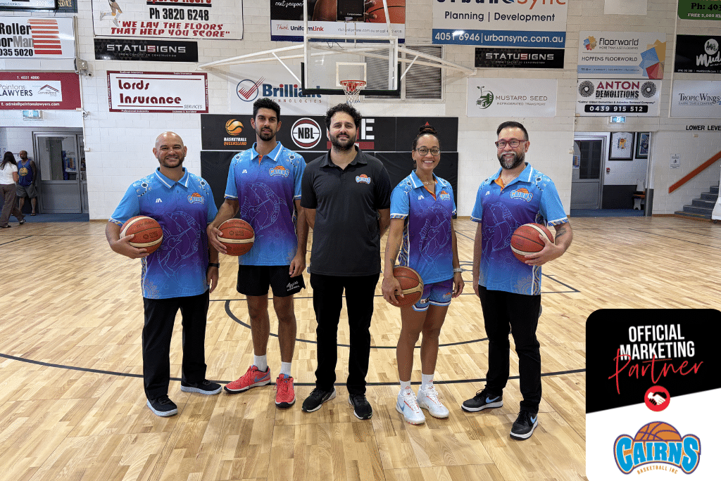 Five people pose on a basketball court in blue uniforms, each holding a basketball for a group photo with sponsor banners in the background and a court backdrop nearby.